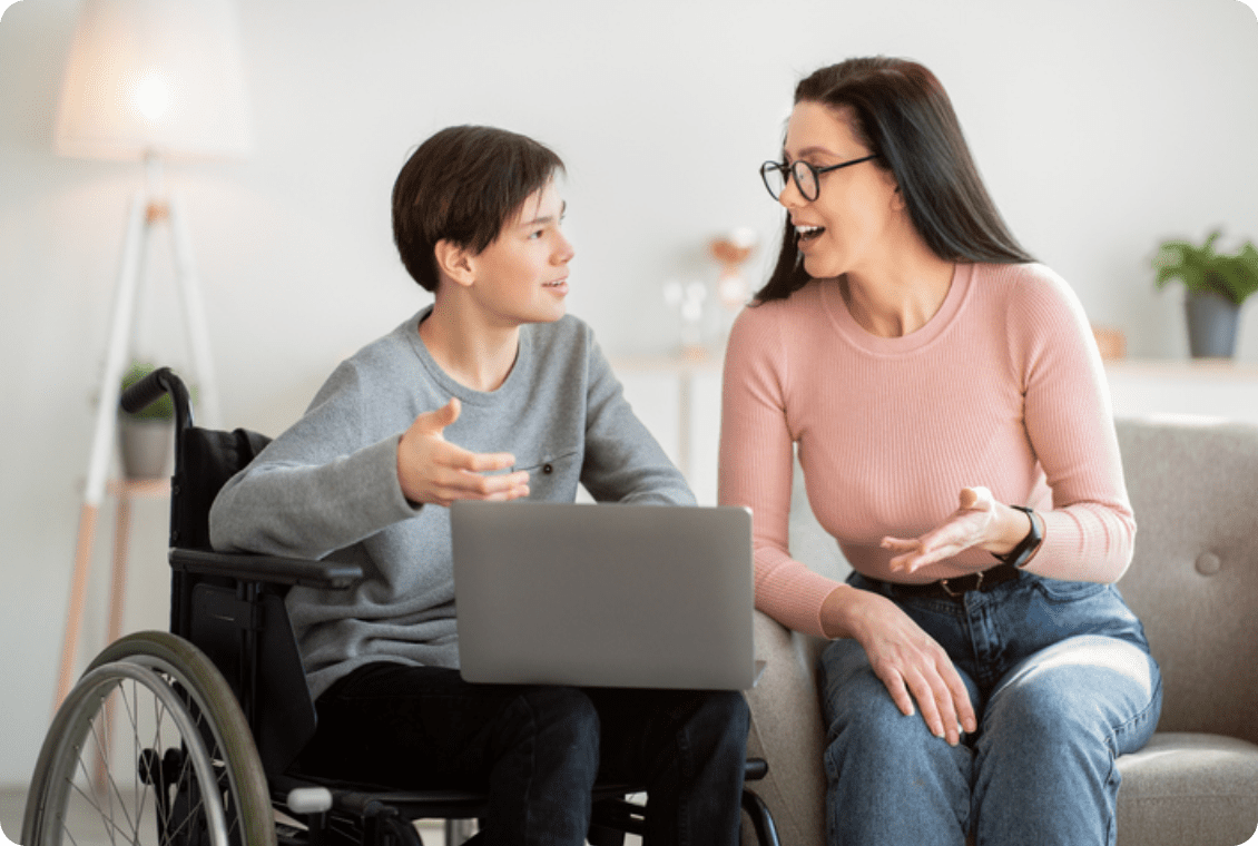 Cheerful disabled teen boy in wheelchair holding laptop, talking to his mom or teacher indoors